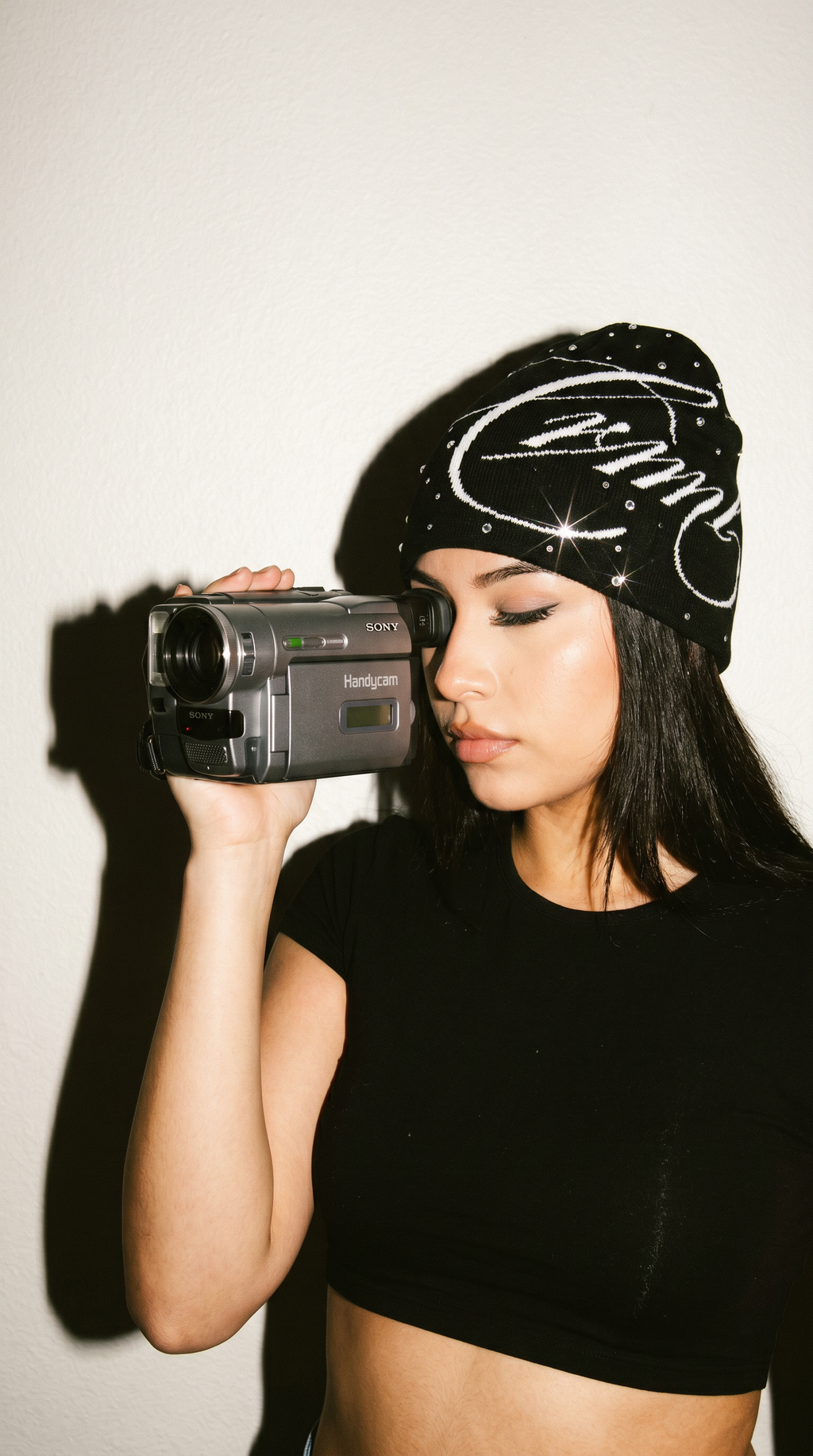 Woman holding a camera with a black beanie and black top against a white background